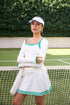 Model wearing a white women’s tennis top with pleated skort and long-sleeve top on an outdoor tennis court.