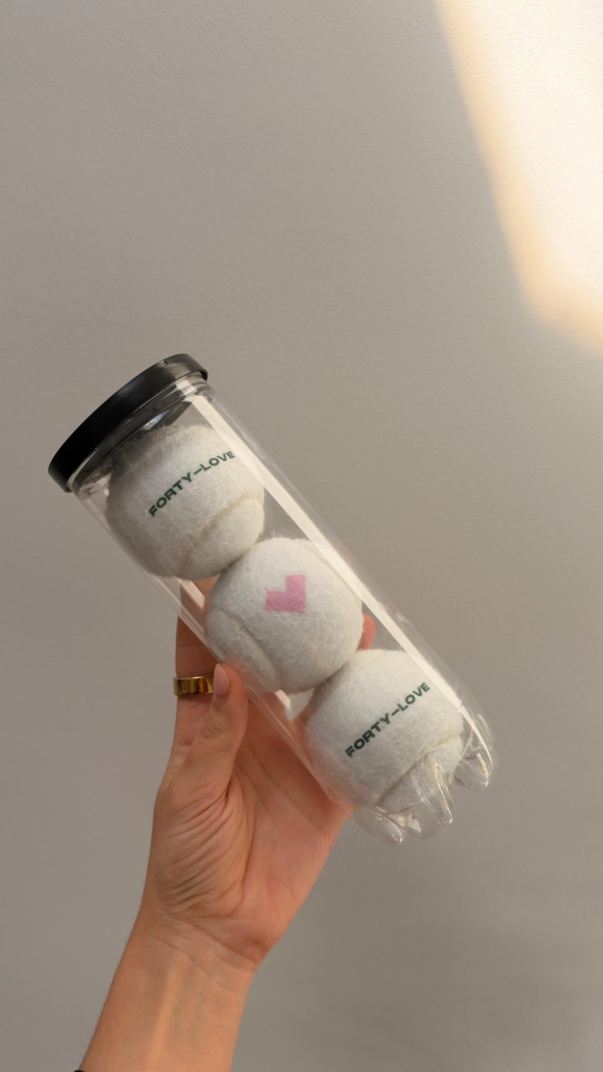 Hand holding a clear Forty-Love can with three white tennis balls: green FORTY-LOVE on the top and bottom balls and a pink heart on the middle ball.