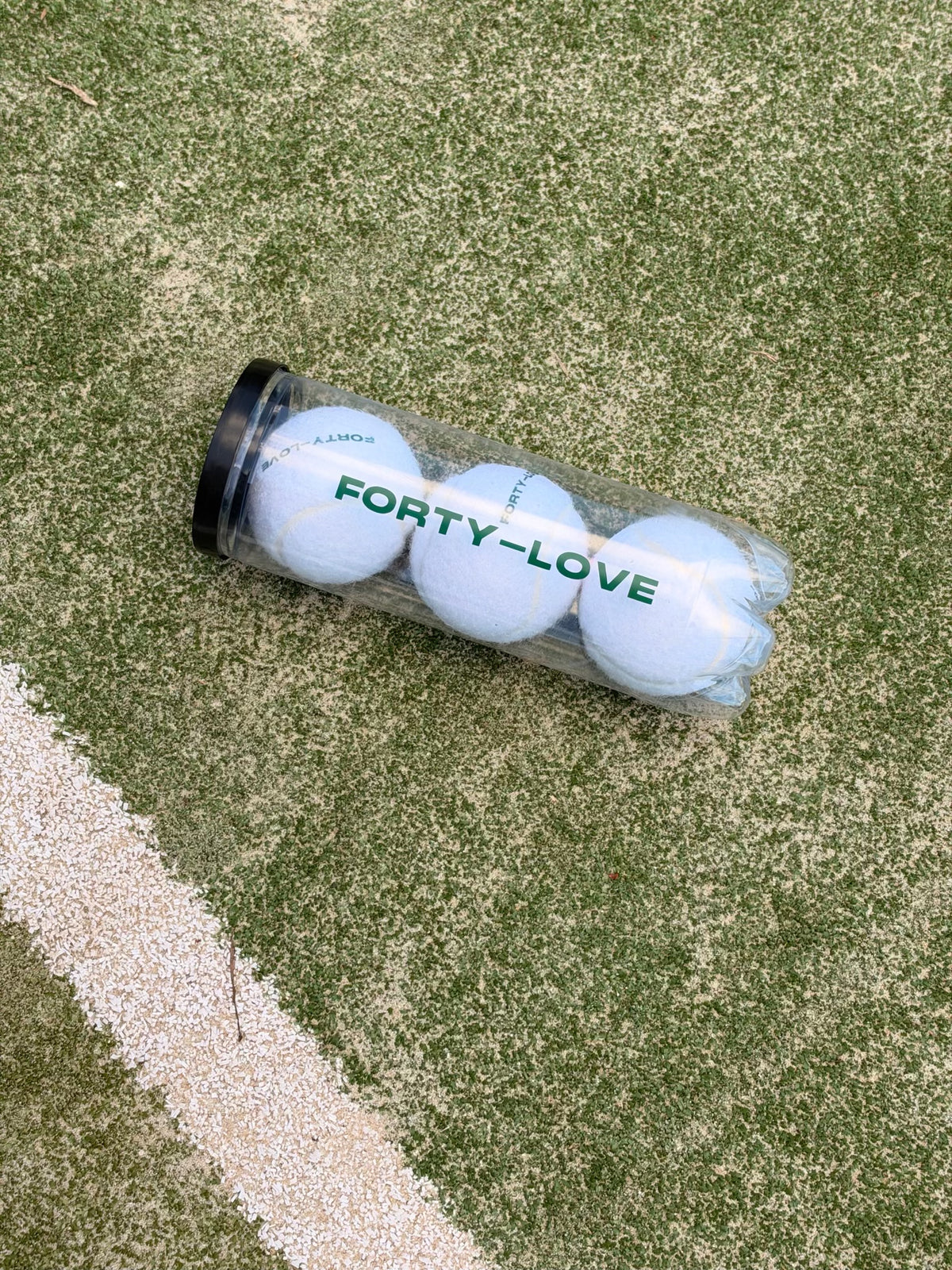 Three white tennis balls in a clear Forty-Love printed can with a black lid, resting beside a white line on a tennis court.