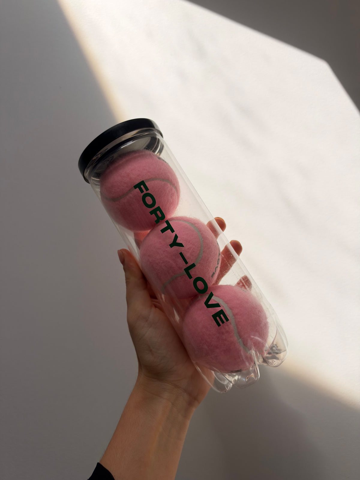 Hand holding three pink tennis balls in a clear Forty-Love printed can with a black lid, in sunlight against a wall.