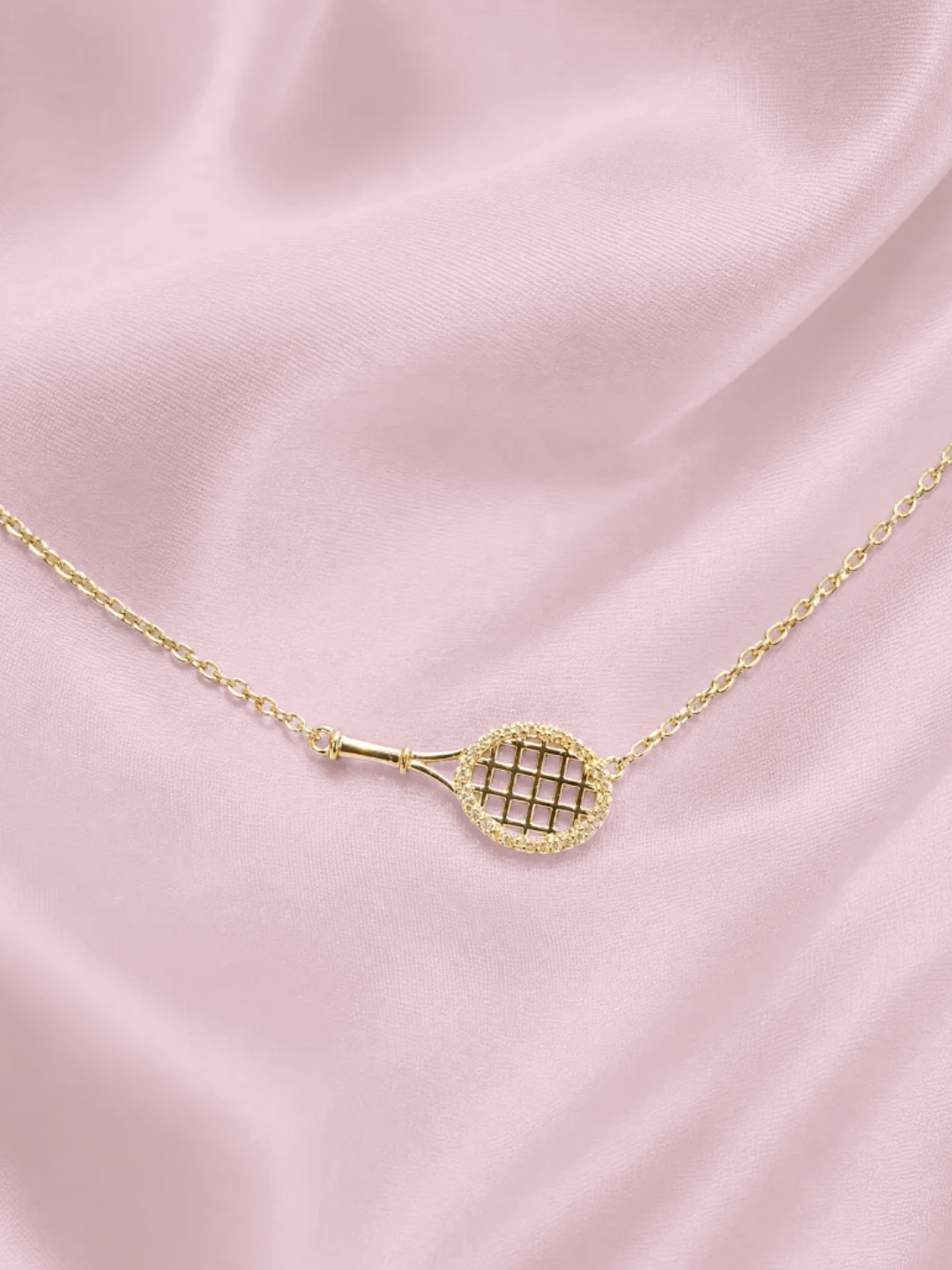 Gold tennis racquet pendant necklace with a pave racquet head, resting on pink satin