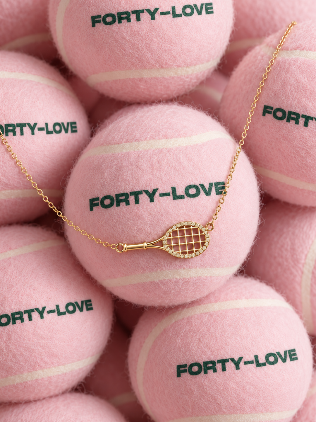 Gold tennis racquet pendant necklace draped over pink Forty-Love tennis balls