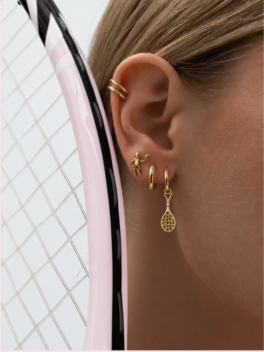 Gold tennis racquet charm hoop earring worn with stacked gold hoops against a pink racquet frame