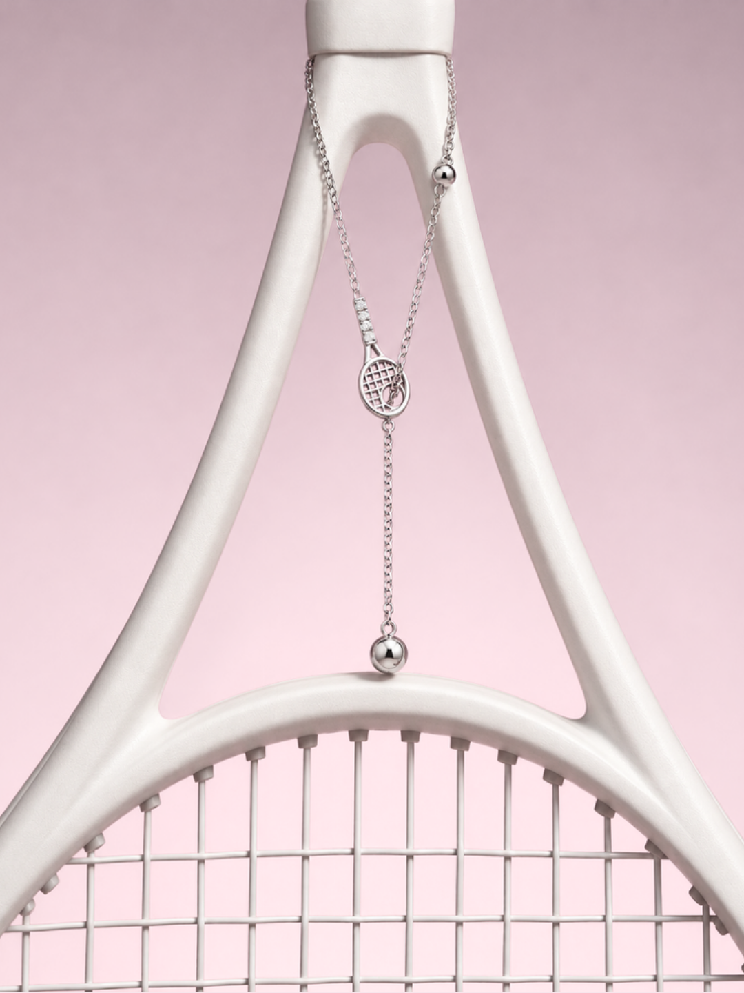 Silver lariat necklace with a tennis racquet charm and ball drops, hanging from a white racquet frame against pink