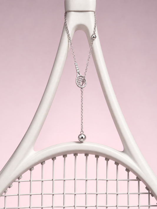 Silver lariat necklace with a tennis racquet charm and ball drops, hanging from a white racquet frame against pink