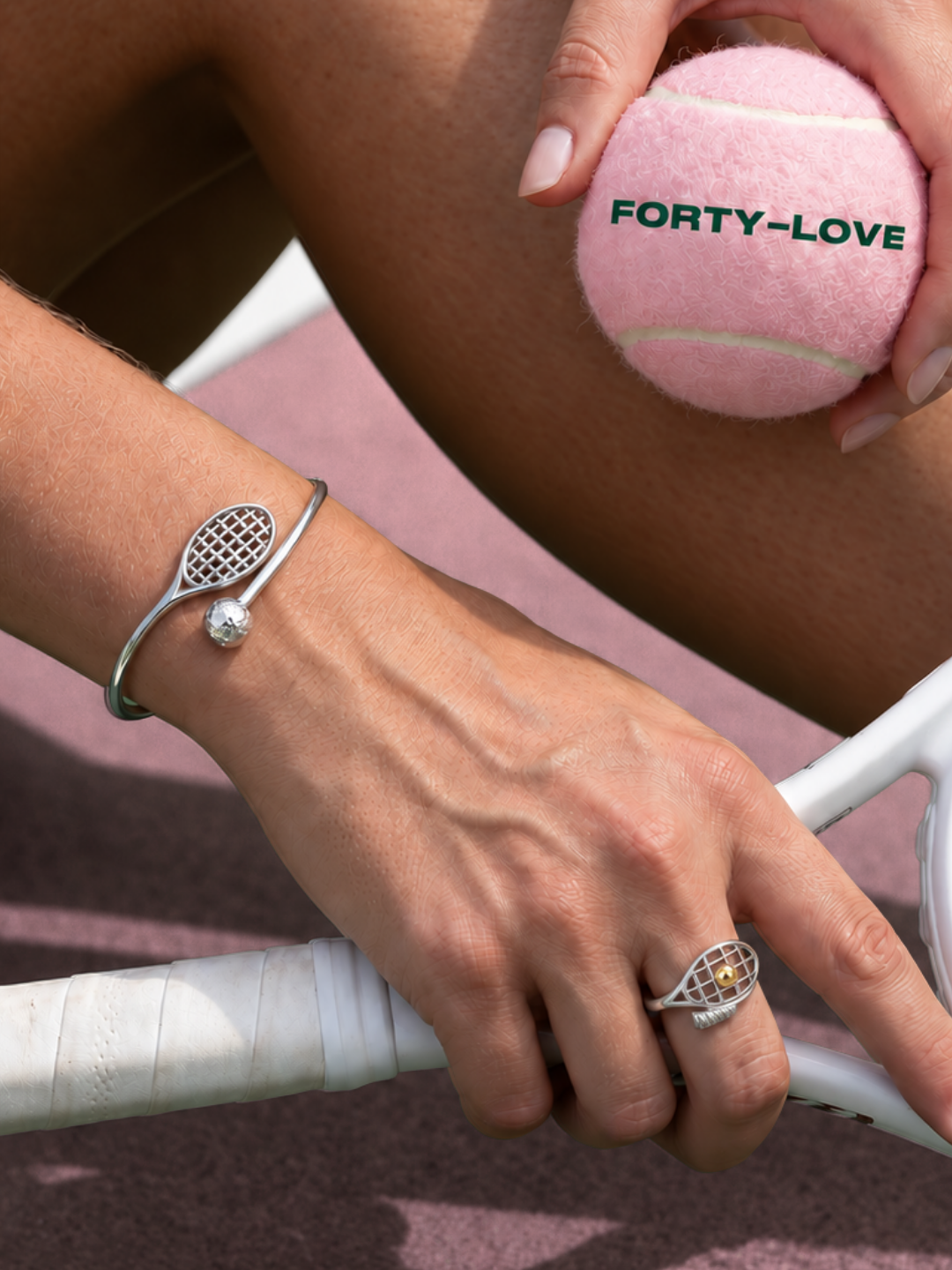 Silver tennis racquet bangle bracelet and racquet ring worn on a hand gripping a racquet, holding a pink Forty-Love tennis ball